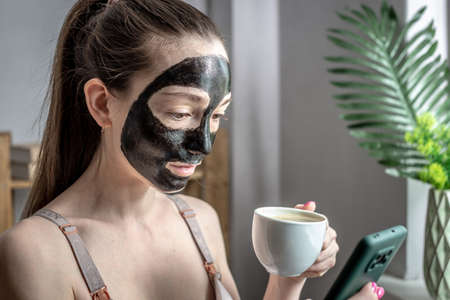 Woman with a cosmetic black mask on her face with a cup of coffee in her hand is using a mobile phone. Concept of lifestyle, beauty treatments, leisure and relaxation.の写真素材