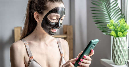 Woman with a cosmetic black mask on her faceis using a mobile phone. Concept of lifestyle, beauty procedures, leisure and relaxation.の写真素材
