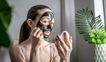 Young woman is holding a mirror in her hand and removing a cosmetic black mask from her face. Concept of skin care, beauty treatment process at home, lifestyle.の写真素材