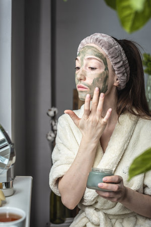 Young beautiful woman in a dressing bathrobe is applying a green cosmetic mask to her face. Concept of skin care and lifestyle.の写真素材