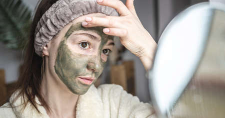 Young pretty woman in a bathrobe in front of a mirror is applying a green cosmetic mask to her face. Concept of skin care, the use of natural cosmetics and lifestyle.の写真素材
