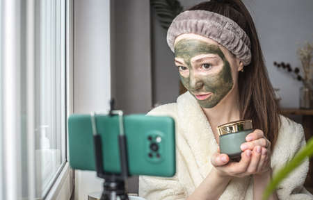 Pretty blogger woman in a bathrobe with a green cosmetic mask on her face is showing a jar of mask on a mobile phone camera.の写真素材