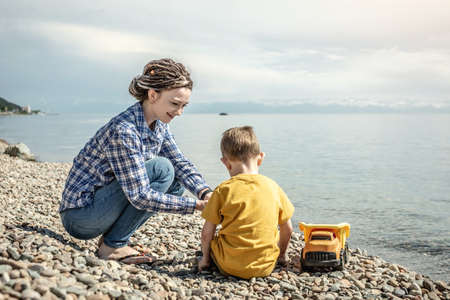 Happy mother and little son on the shore of a large pond are playing together and collecting stones....