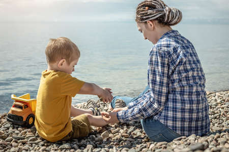 Mom and little child on the stony coast of a large lake are playing together and collecting stones. Concept of family leisure, pastime in nature, lifestyle.の写真素材