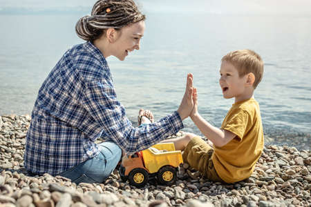 Happy mother and little son on the shore of a large pond are playing together and collecting stones. Concept of family leisure, pastime in nature, lifestyle.の写真素材