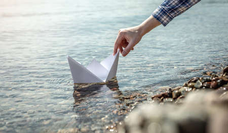 Woman's hand is launching a white paper boat into the clear water of the sea with a stony bottom.の写真素材