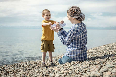 Mom and little child on the stony coast of a large lake are playing together and making paper boats....