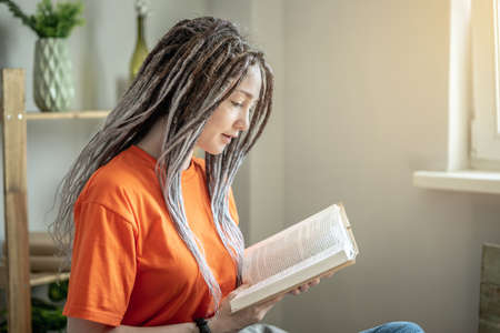 Young woman with dreadlocks and bright clothes is sitting and reading a book at home. Concept of leisure, pastime, learning and lifestyle.の写真素材