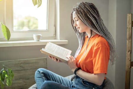 Young woman with dreadlocks and bright clothes is sitting comfortably in a bag chair and reading a...