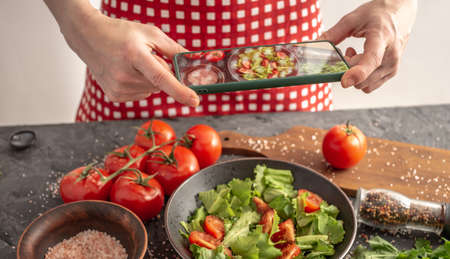 Women's hands are holding a mobile phone over a beautiful appetizing vegetable salad to take a...