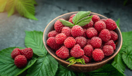 Bowl with fresh ripe delicious raspberries and a green leaves on a black background.