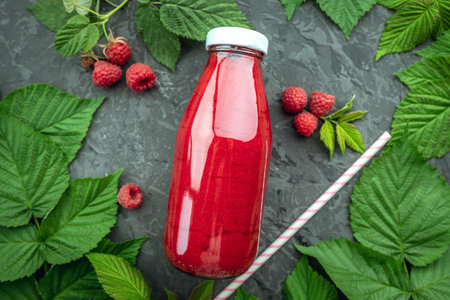 Glass bottle of fresh raspberry smoothie and a straw on a black table covered with raspberry leaves and berries. Concept of freshening and healthy summer drinks. Top view.の写真素材