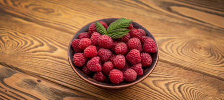 Bowl with fresh ripe delicious raspberries and a green leaf on a wooden background.