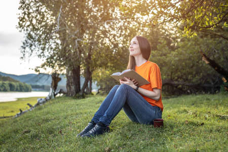 A young beautiful woman in bright clothes is sitting on the green grass and enjoying reading a book in nature. Concept of a pleasant pastime, rest, relaxation.の写真素材