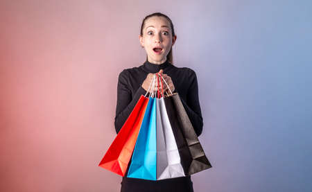 Woman with surprised face in a black dress is holding a colorful paper bags with purchases from a shop in her hands on a blue pink background. Concept of Black Friday, sale and shopping.の写真素材