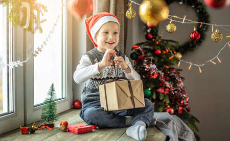 Happy boy in a red Santa hat is sitting at the window next to a Christmas tree in a room decorated for the holiday and holding a gift box in his hands. Concept of New Year atmosphere and festive mood.の写真素材