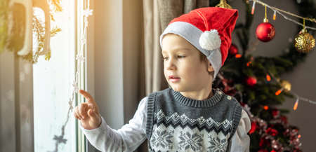 Cute little boy in a red Santa hat is sitting at the window next to the Christmas tree and looking at the festive decorations at home. Concept of New Year atmosphere and holiday mood.の写真素材