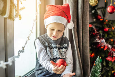 Cute little boy in a Santa hat is sitting next to a Christmas tree in a decorated room and holding a Christmas toy in his hands. Concept of preparing for the New Year, the atmosphere of the holiday.の写真素材