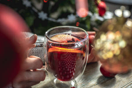 Woman in a sweater is holding a clear mug with fragrant spicy mulled wine decorated with fruits and...