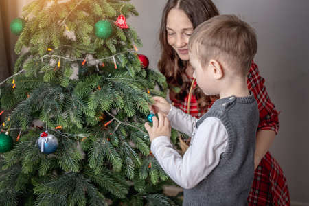Mom with her child is decorating together the Christmas tree with toys and balls. Concept of family preparation for the New Year, cozy atmosphere and festive mood.の写真素材