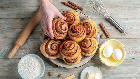 Hand is gently picking up a fresh cinnamon roll from a plate full of buns. Concept of delicious...