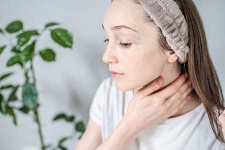 Young woman on the background of a green plant is doing a massage and smearing her neck with a natural organic cream or oil. Concept of spending time alone, relaxing, skin care and self care.の写真素材