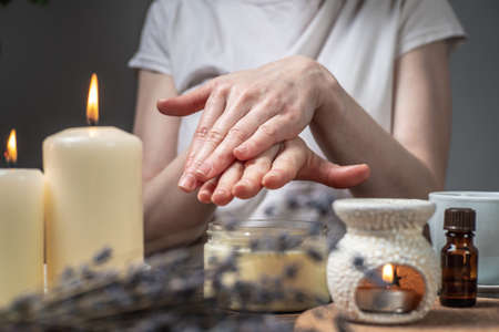 Woman is smearing her hands with a natural organic cream doing a massage. Aroma lamp with essential oils and candles on the table. Concept of skin and self care, atmosphere of harmony and relaxationの写真素材