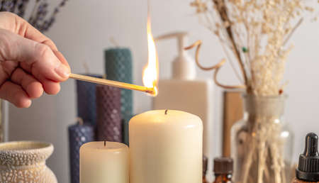 Hand is lighting the candle with a burning match. There is aroma lamp, candles and cosmetics on the table. Concept of aromatherapy, relaxing atmosphere.の写真素材