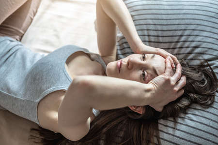 Young woman is lying in bed and holding her head with her hands experiencing a headache, migraine. Concept of insomnia caused by stress, anxiety and problems.の写真素材