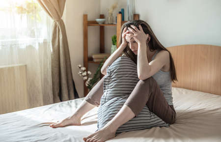 Young woman is sitting in bed and holding her head with her hands experiencing a headache, migraine. Concept of insomnia caused by stress, problems and anxious thoughts.の写真素材