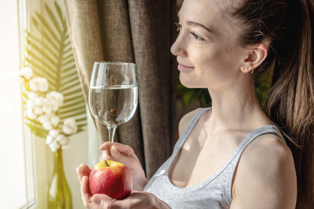 Young woman is standing in front of the window on a sunny morning and holding a glass of clear water and an apple in her hands. Concept of a pleasant start to the day and healthy habits.の写真素材