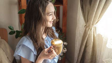 Happy cute woman with a cup of aromatic coffee in her hands is smiling and looking out the window on...