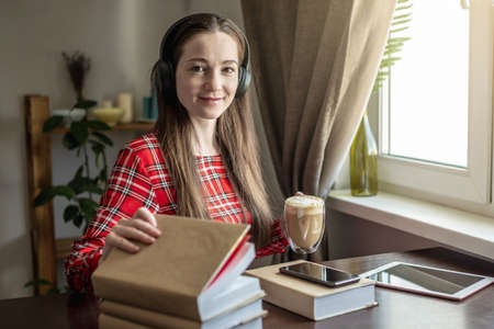 A woman in a red dress is listening audiobook with headphones and drinking coffee by the window....