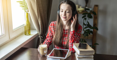 A woman in a red dress is listening audiobook wearing headphones with tablet by the window. The...
