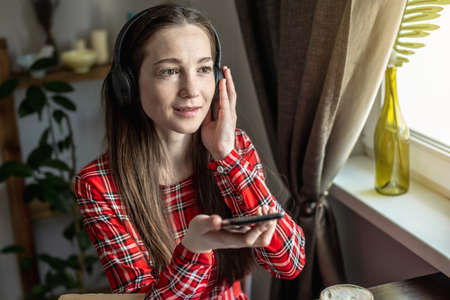 A woman in a red dress is listening audiobook wearing headphones with phone by the window. The...