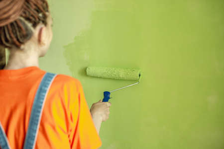 Woman with dreadlocks, in an orange t-shirt and denim jumpsuit is painting the wall with a roller in green color. Concept of repair, renovation of the new apartment. copyspace.の写真素材