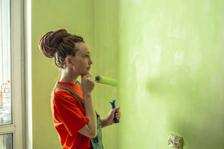 Woman with dreadlocks, in an orange t-shirt and denim jumpsuit is painting the wall with a roller in...