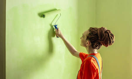 Woman with dreadlocks, in an orange t-shirt and denim jumpsuit is painting the wall with a roller in green color. Concept of repair, renovation of the new apartment. copyspace.の写真素材