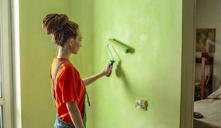 Woman with dreadlocks, in an orange t-shirt and denim jumpsuit is painting the wall with a roller in green color. Concept of repair, renovation of the new apartment. copyspace.の写真素材