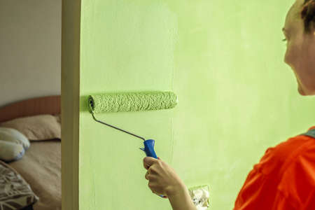 Woman with dreadlocks, in a bright orange t-shirt is painting the wall with a roller in green color. Concept of repair, renovation of the new apartment.の写真素材