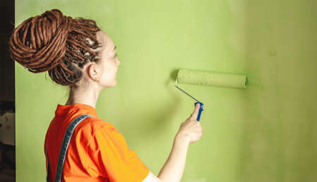 Woman with dreadlocks, in an orange t-shirt and denim jumpsuit is painting the wall with a roller in green color. Concept of renovation, renovation of the new apartmentの写真素材