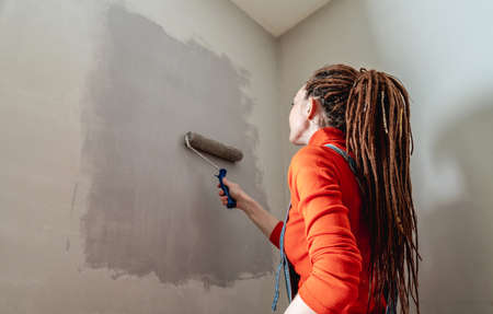 Woman with dreadlocks, in an orange turtleneck and denim jumpsuit is painting the wall with a roller...