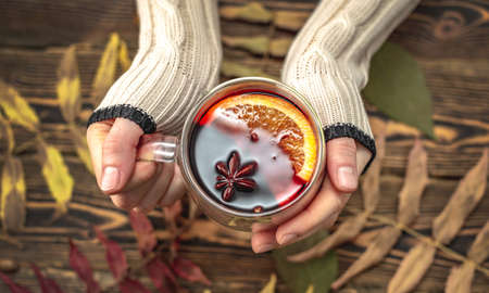 Hands of woman in a sweater are holding a glass cup with mulled wine on a wooden background with autumn leaves. Concept of a traditional warming beverage.の写真素材