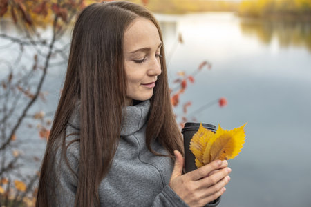 Woman is walking through the autumn forest along the river with a cup of coffee and enjoying the beautiful nature, fresh air and calm.の写真素材