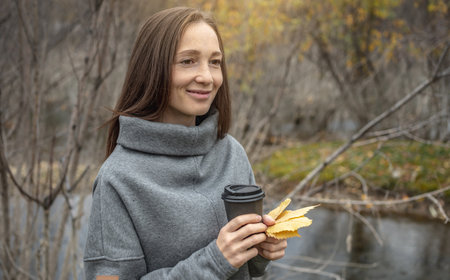 Woman is walking through the autumn forest along the river with a cup of coffee and enjoying the beautiful nature, fresh air and calm.の写真素材