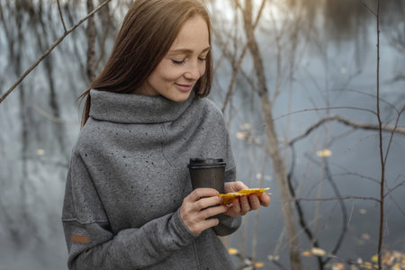 Woman is walking through the autumn forest along the river with a cup of coffee and enjoying the beautiful nature, fresh air and calm.の写真素材