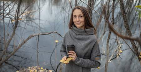 Young woman with a cup of coffee and a bouquet of autumn leaves in the forest near a pond in a fine rain.の写真素材