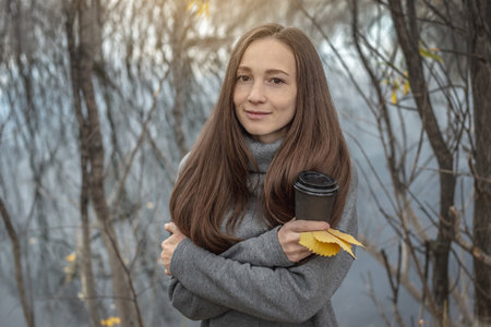 Young woman is walking through a beautiful autumn forest along the river and warming up with hot coffee.の写真素材