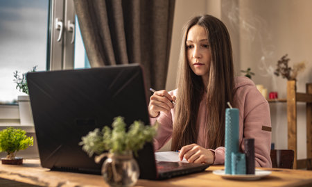 Woman is sitting at a table in front of a laptop, listening attentively and writing down information in a notebook. Concept of distance learning and video communication.の写真素材