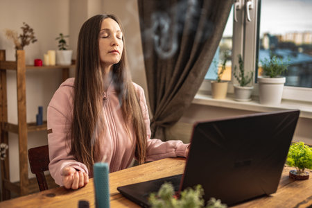 Woman is sitting at a table in front of a laptop, meditating and relaxing while listening to an instructor online or a video lecture. Concept of a remote therapy and meditation session.の写真素材